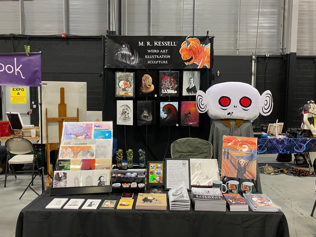 M. R. Kessell's vendor display of paintings, prints, books, and his life-sized Batboy costume at the September 2022 Trenton Punk Rock Flea Market